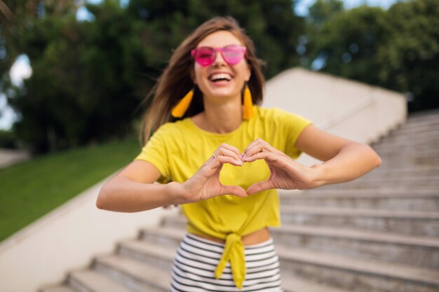 Ritratto di giovane donna abbastanza sorridente che si diverte nel parco cittadino, positivo, emotivo, indossa un top giallo, orecchini, occhiali da sole rosa, tendenza moda stile estivo, accessori alla moda, mostrando il segno del cuore