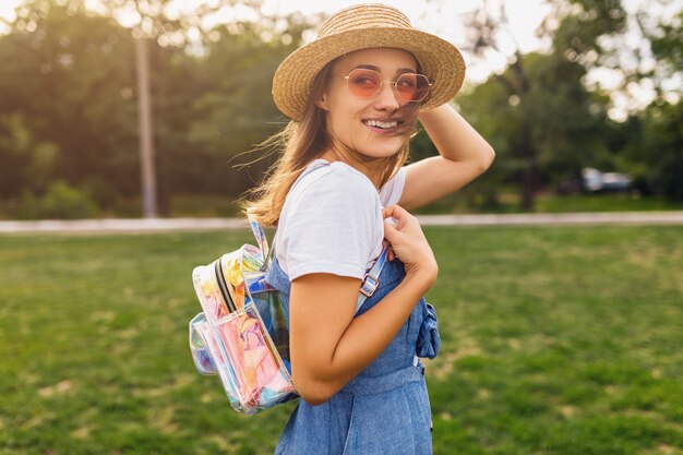 Ritratto di giovane donna abbastanza sorridente in cappello di paglia e occhiali da sole rosa che cammina nel parco, stile di moda estiva, vestito colorato hipster