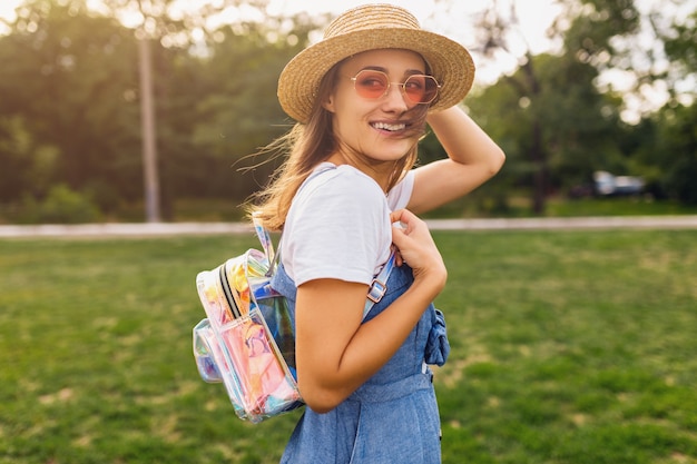 Ritratto di giovane donna abbastanza sorridente in cappello di paglia e occhiali da sole rosa che cammina nel parco, stile di moda estiva, vestito colorato hipster