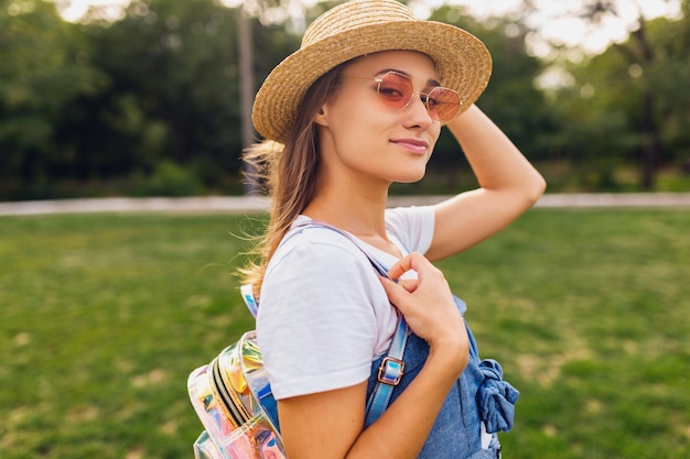 Ritratto di giovane donna abbastanza sorridente in cappello di paglia e occhiali da sole rosa che cammina nel parco, stile di moda estiva, vestito colorato hipster