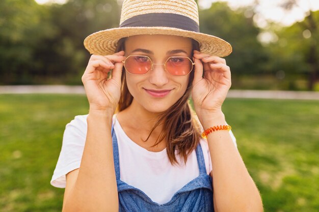 Ritratto di giovane donna abbastanza sorridente in cappello di paglia e occhiali da sole rosa che cammina nel parco, stile di moda estiva, vestito colorato hipster