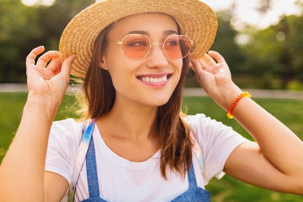 Ritratto di giovane donna abbastanza sorridente in cappello di paglia e occhiali da sole rosa che cammina nel parco, stile di moda estiva, vestito colorato hipster