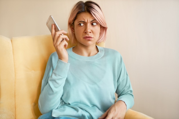 Ritratto di giovane donna accigliata in felpa blu che tiene mobile, componendo un numero sbagliato, avendo uno sguardo scioccato.