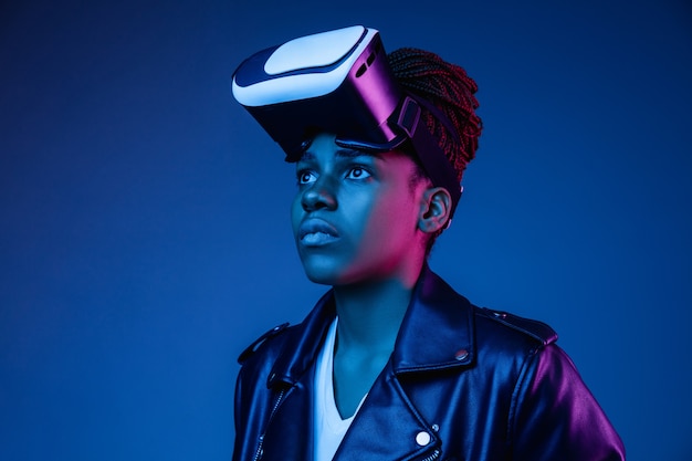 Ritratto di giovane donna afro-americana che gioca in occhiali VR in luce al neon sull'azzurro.