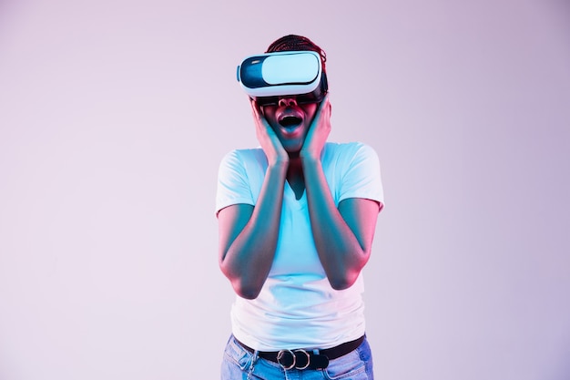 Ritratto di giovane donna afro-americana sta giocando in occhiali VR in luce al neon su sfondo sfumato. Concetto di emozioni umane, espressione facciale, gadget moderni e tecnologie. Guarda stupito.