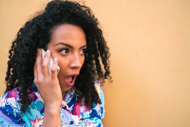 Ritratto di giovane donna afro parlando al telefono con espressione scioccata su sfondo giallo. Concetto di comunicazione.