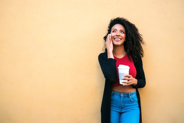 Ritratto di giovane donna afro parlando al telefono mentre si tiene una tazza di caffè su sfondo giallo. Concetto di comunicazione.