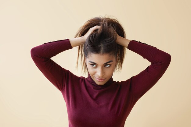 Ritratto di giovane donna afroamericana elegante in maglione dolcevita alla moda che guarda lontano con un sorriso misterioso come se avesse una grande idea mentre si fa i capelli, preparandosi per il lavoro la mattina