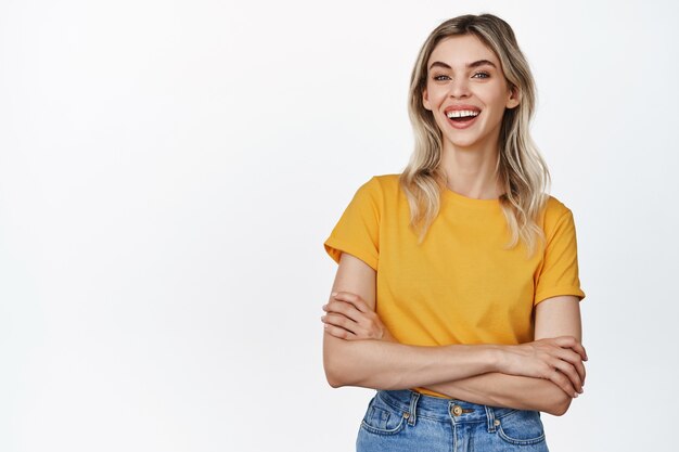 Ritratto di giovane donna alla moda con capelli biondi su sorriso bianco, braccia incrociate sul petto e ridendo, in piedi in maglietta gialla su bianco.