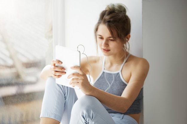 Ritratto di giovane donna allegra adolescente in cuffie sorridenti guardando tablet navigazione web navigazione internet seduto vicino alla finestra sul muro bianco.