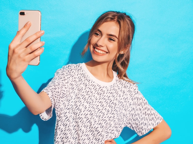 Ritratto di giovane donna allegra che prende il selfie della foto. Bella ragazza che tiene la fotocamera dello smartphone. Posa di modello sorridente vicino alla parete blu in studio