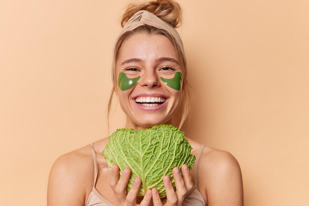 Ritratto di giovane donna allegra utilizza cosmetici naturali e prodotti di bellezza tiene cavolo fresco verde per mettere la pelle per idratare applica cerotti idrogel sotto gli occhi si erge spalle nude