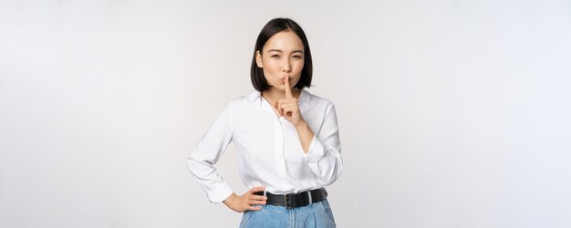 Ritratto di giovane donna asiatica adulta shushing dire silenzio shh premere il dito sulle labbra condividendo il segreto non parlare gesto tabù in piedi su sfondo bianco