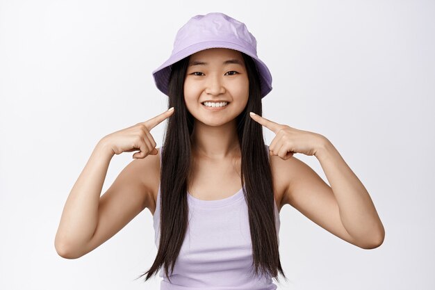 Ritratto di giovane donna asiatica con cappello a secchiello, puntando il dito contro il suo sorriso bianco, concetto di clinica odontoiatrica e sbiancamento dei denti, studio