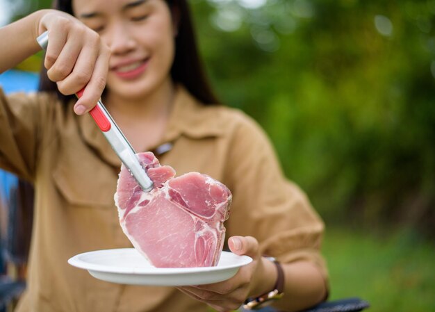 Ritratto di giovane donna asiatica felice che si accampa da solo barbecue di maiale alla griglia nella padella da picnic e cucina il cibo mentre si siede su una sedia nel campeggio