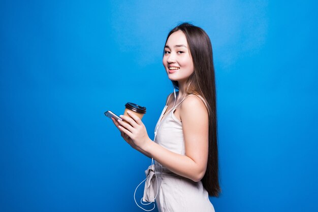 Ritratto di giovane donna asiatica in piedi sorriso, utilizzando il telefono cellulare la sua azienda tazza di carta da caffè, guardando smartphone sulla parete blu