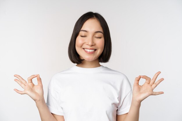 Ritratto di giovane donna asiatica meditando sorridente soddisfatto e pratica yoga chiudere gli occhi e meditare respiro calmo in piedi su sfondo bianco