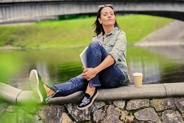 Ritratto di giovane donna attraente beve caffè in movimento in un parco estivo vicino al lago.