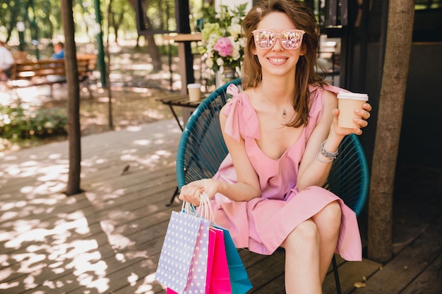 Ritratto di giovane donna attraente felice sorridente che si siede nella caffetteria con borse della spesa bere caffè, vestito di moda estate, abito di cotone rosa, abbigliamento alla moda