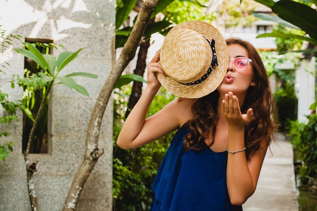 Ritratto di giovane donna attraente felice sorridente in vestito blu e cappello di paglia che indossa occhiali da sole rosa camminando in hotel villa spa tropicale in vacanza in abito stile estivo