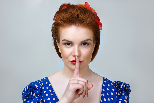 Ritratto di giovane donna attraente in abito retrò che nasconde informazioni segrete o riservate, tenendo il dito indice sulle labbra rosse