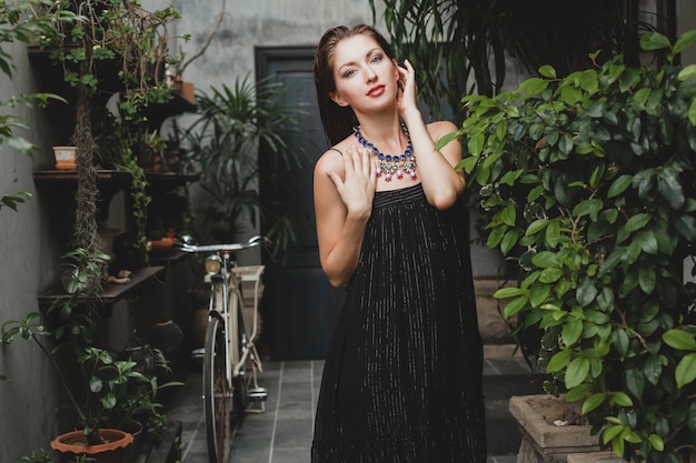 Ritratto di giovane donna attraente in elegante abito nero in posa in villa tropicale, sexy, stile estivo elegante, accessori alla moda collana, sorridente, gioielli, pura pelle naturale del viso