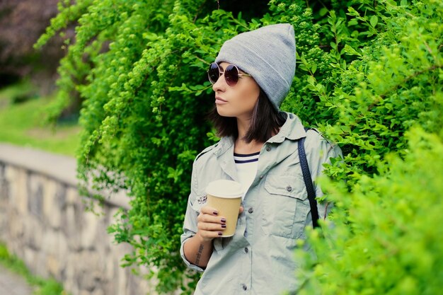 Ritratto di giovane donna attraente in occhiali da sole tiene una tazza di caffè di carta in un verde parco estivo.