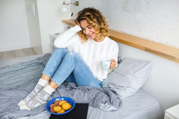 Ritratto di giovane donna attraente seduta sul letto la mattina, bere il caffè in tazza, mangiare i biscotti