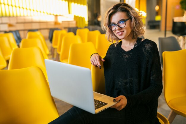 Ritratto di giovane donna attraente seduto in aula, lavorando sul portatile, con gli occhiali, aula, molte sedie gialle, formazione degli studenti online, libero professionista, elegante