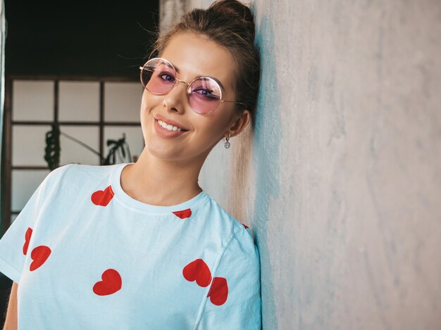 Ritratto di giovane donna bellissima, guardando la fotocamera Ragazza alla moda in casual estate t-shirt bianca e occhiali da sole Donna divertente e positiva in posa vicino al muro grigio in studio