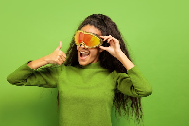 Ritratto di giovane donna bruna afro-americana in passamontagna su sfondo verde studio. Concetto di emozioni umane, espressione facciale, vendite, pubblicità, sport invernali e vacanze. Sorridendo, pollice in su.