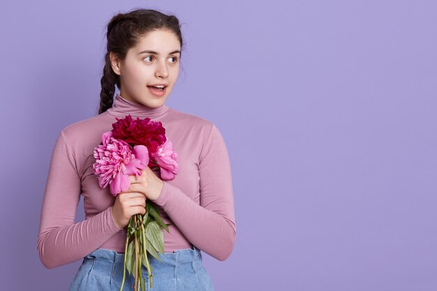 Ritratto di giovane donna bruna che indossa abbigliamento casual, abbracciando bouquet di peonie, donna che osserva da parte, in piedi contro il muro lilla con la bocca aperta, stupito.