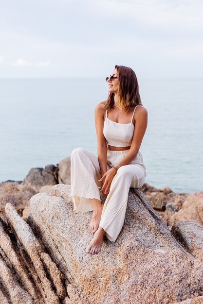 Ritratto di giovane donna calmo felice indoeuropeo in forma slim fit in top e pantaloni crop cami si siede da solo sulla spiaggia tropicale rocciosa al tramonto