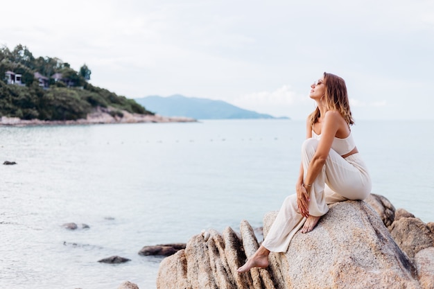 Ritratto di giovane donna calmo felice indoeuropeo in forma slim fit in top e pantaloni crop cami si siede da solo sulla spiaggia tropicale rocciosa al tramonto