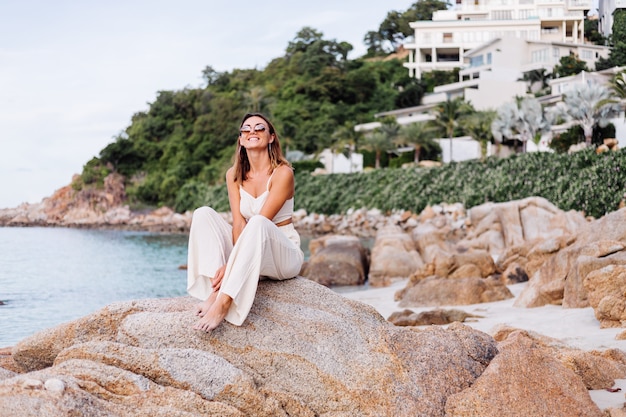 Ritratto di giovane donna calmo felice indoeuropeo in forma slim fit in top e pantaloni crop cami si siede da solo sulla spiaggia tropicale rocciosa al tramonto