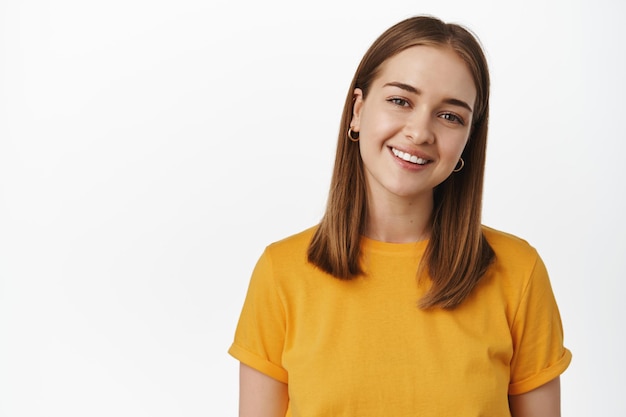 Ritratto di giovane donna candida che sorride denti bianchi, inclina la testa amichevole e guarda felice alla telecamera, indossa una t-shirt gialla su sfondo bianco. Copia spazio