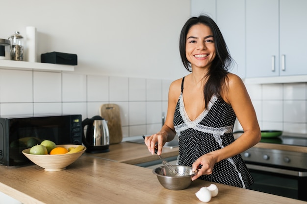 Ritratto di giovane donna castana graziosa che cucina uova strapazzate in cucina al mattino, sorridente, umore felice, casalinga positiva, stile di vita sano