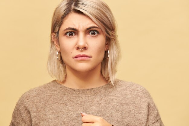 Ritratto di giovane donna caucasica arrabbiata lunatica alla moda che indossa piercing facciale e maglione caldo che aggrotta le sopracciglia di cattivo umore, dimostrando la sua disapprovazione e malcontento. Emozioni negative