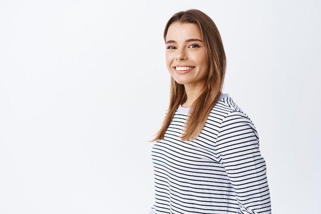 Ritratto di giovane donna caucasica con capelli biondi e sorriso bianco, gira la testa davanti e sembra amichevole e naturale davanti, in piedi sul muro bianco