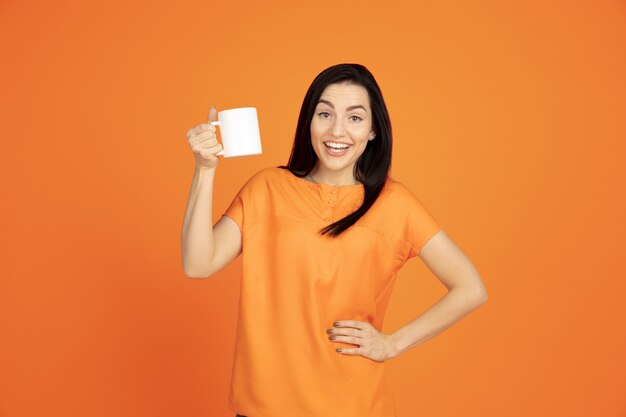 Ritratto di giovane donna caucasica su sfondo arancione studio. Bello modello femminile del brunette in camicia. Concetto di emozioni umane, espressione facciale, vendite, annuncio. Copyspace. Bere caffè o tè.