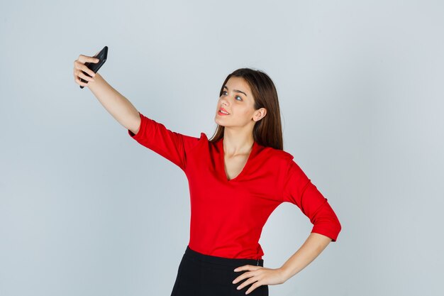 Ritratto di giovane donna che cattura selfie mantenendo la mano sul fianco in camicetta rossa