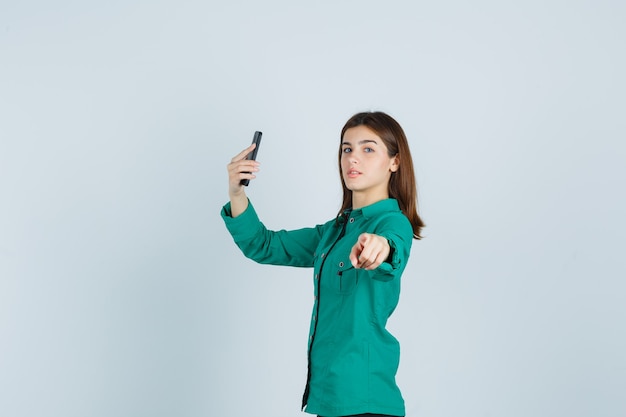Ritratto di giovane donna che cattura selfie sul telefono cellulare mentre punta alla telecamera in maglietta verde e guardando fiducioso