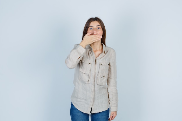 Ritratto di giovane donna che copre la bocca con la mano in casual, jeans e sembra scioccata vista frontale