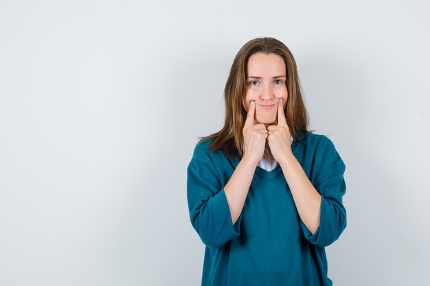 Ritratto di giovane donna che costringe un sorriso sul viso con le dita in camicia bianca, maglione e guardando la vista frontale dispiaciuta