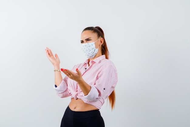 Ritratto di giovane donna che finge di mostrare qualcosa in camicia, pantaloni, maschera e guardando perplesso vista frontale