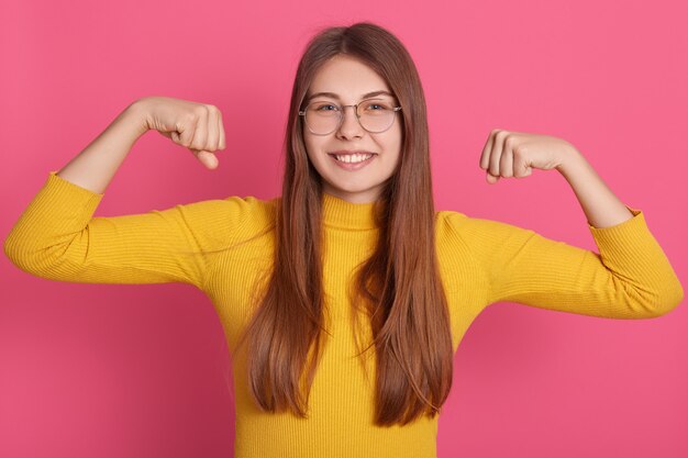 Ritratto di giovane donna che flette i muscoli e che sorride contro la parete ottimistica. Modello femminile europeo in abbigliamento casual che mostra i suoi muscoli