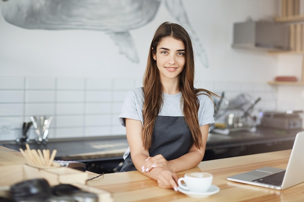 Ritratto di giovane donna che gestisce un caffè di successo che beve cappuccino appena fatto e usa un computer portatile