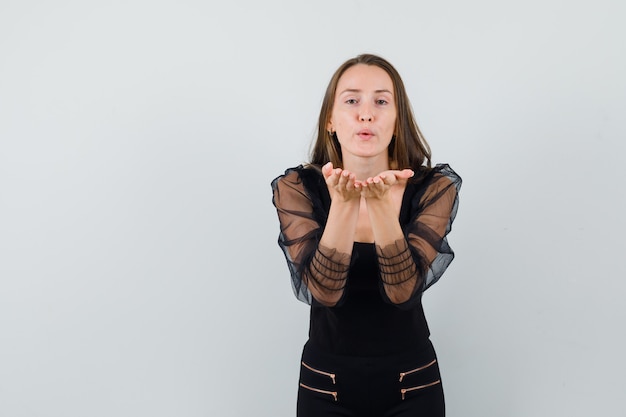 Ritratto di giovane donna che invia baci alla telecamera in camicetta nera e pantaloni neri e guardando grave vista frontale