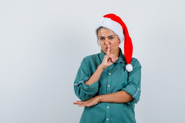 Ritratto di giovane donna che mostra gesto di silenzio in camicia, cappello da Babbo Natale e guardando fiducioso vista frontale