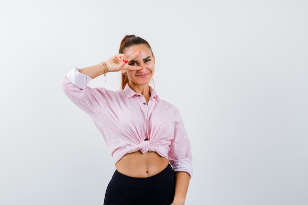 Ritratto di giovane donna che mostra il segno a V sull'occhio in camicia, pantaloni e vista frontale allegra